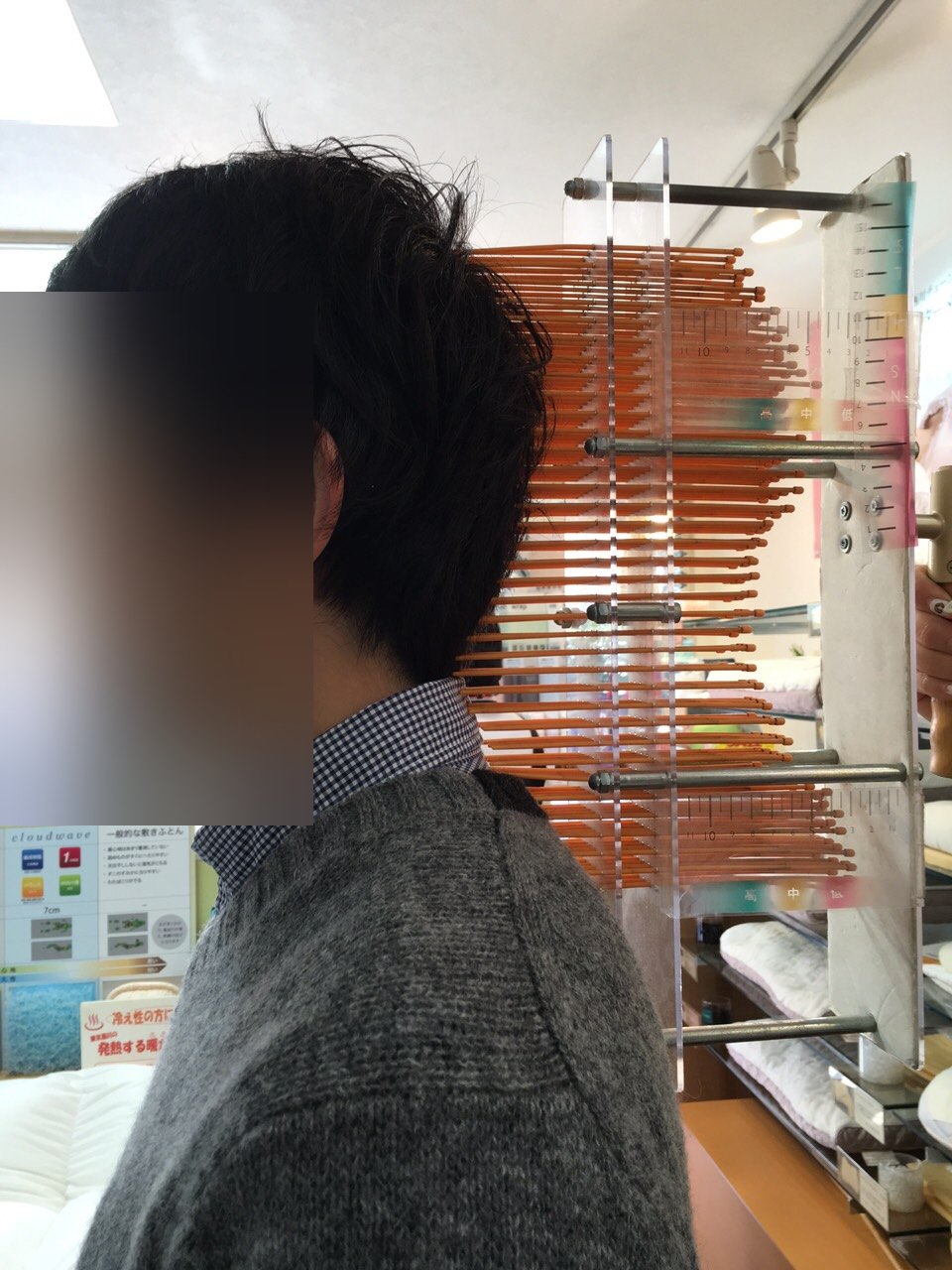 寝具店で、首のうしろに細い棒がたくさん並んだ器具を当てて、首のカーブを測っているところ